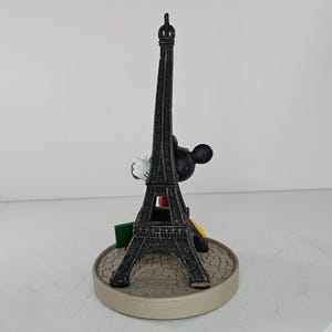 K&ouml;nnte beinhalten: Dekorative Figur mit dem Eiffelturm und Mickey Mouse. Der Turm ist dunkelbraun, Mickey Mouse ist schwarz mit wei&szlig;en Handschuhen. Der Sockel ist hellgrau mit einem Kopfsteinpflastermuster. Ein Souvenir.