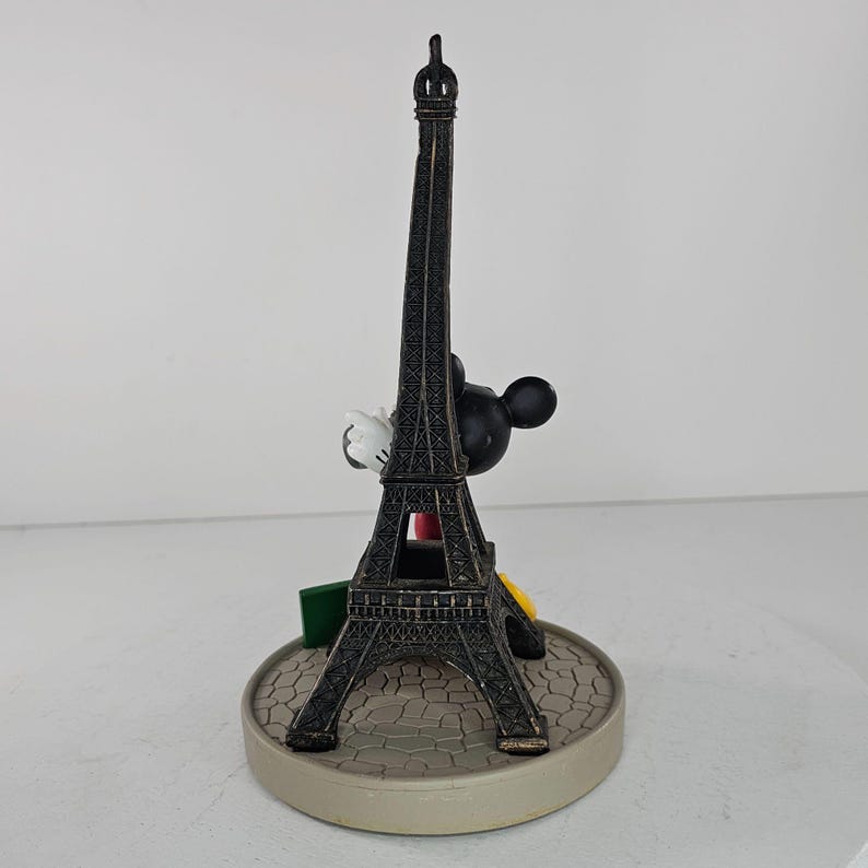 K&ouml;nnte beinhalten: Dekorative Figur mit dem Eiffelturm und Mickey Mouse. Der Turm ist dunkelbraun, Mickey Mouse ist schwarz mit wei&szlig;en Handschuhen. Der Sockel ist hellgrau mit einem Kopfsteinpflastermuster. Ein Souvenir.