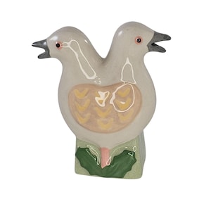 May include: A ceramic sculpture of a two-headed bird. The bird is primarily light gray with a cream-colored chest featuring a yellow and cream design. The beaks are black, and the eyes are red. The base is green with a pink and green leaf design.
