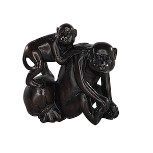 Puede incluir: Una escultura marrón oscuro de dos monos. Uno está sentado encima de un objeto redondo, mientras que el otro está sobre la espalda del primero. La escultura está sobre un fondo blanco.