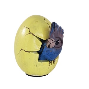 May include: A yellow ceramic egg with a crack down the side, revealing a blue and purple bird figurine inside. The bird has a brown beak and black eyes.