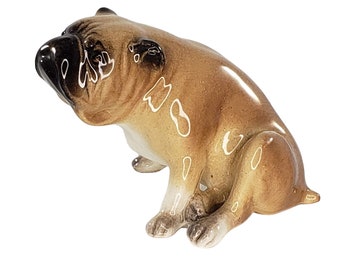 May include: A ceramic figurine of a brown and white bulldog sitting with its head tilted to the side. The figurine has a detailed face and body with white markings.