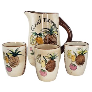May include: A ceramic pitcher and three matching cups with a fruit design. The pitcher has a brown handle and says "Good Morning" on the front. The cups and pitcher feature a pineapple, lemon, and tomato design.