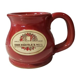Op de afbeelding: Een rode keramische mok met een afgeronde body en een grote handgreep. Een ovaal etiket op de voorkant leest "THE BROKEN MUG" met de tekst "Psalms 147:3". De mok is ontworpen voor warme dranken.