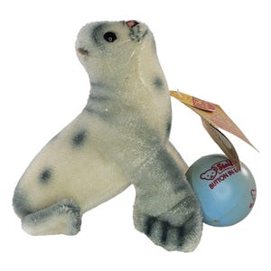 Puede incluir: Un juguete de peluche Steiff vintage con forma de foca en tonos grises y blancos, con manchas grises oscuras. La foca tiene un pequeño ojo negro y una etiqueta adjunta a una bola azul claro. La etiqueta dice "Steiff BUTTON IN EAR".