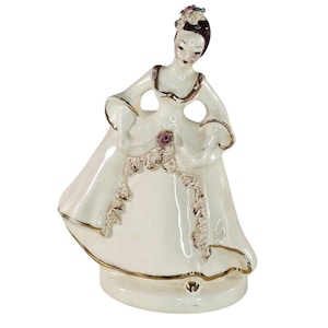 May include: A ceramic figurine of a person wearing a white gown with gold trim, a dark brown hairstyle, and a small floral accent on the head. The dress features a textured lace-like detail down the front and a small pink rose at the waist.