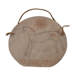Op de afbeelding: Een ronde, beige keramische wandhanger met een bruin leren handvat. Het ontwerp toont een paard, zon en hand symbool in bruin, samen met andere geometrische vormen. Het stuk heeft een rustieke, handgemaakte uitstraling.