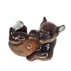 May include: A ceramic figurine of a brown bear cub lying on its back. The bear has a glossy finish with lighter brown accents on its belly and paws. The ears and paws are a lighter shade of brown.