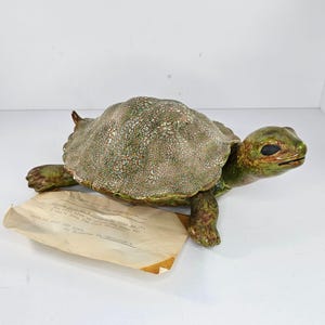 May include: A ceramic turtle figurine with a textured, mottled green and brown shell. The head and legs are a solid green color. The figurine has black eyes and a small, open mouth. A piece of paper with handwritten text rests in front of the turtle.