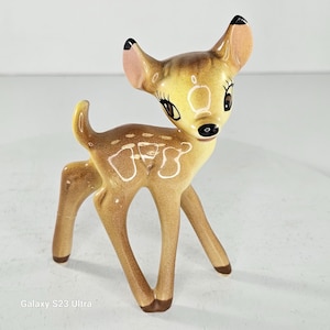Könnte beinhalten: Keramikfigur eines Rehs mit hellbraunem Fell und weißen Flecken. Das Reh hat große, ausdrucksstarke Augen, rosa Ohren und schwarzspitzige Ohren. Die Figur steht auf vier Beinen.