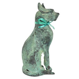 May include: A weathered, seated dog statue with a teal ribbon tied around its neck. The statue has a verdigris finish, giving it a vintage appearance. The dog is depicted in a seated position, with its ears perked up.