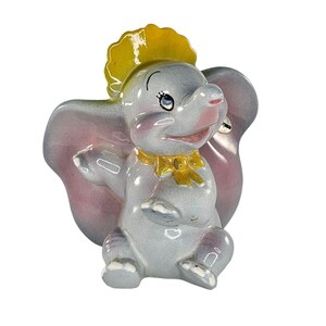 Könnte beinhalten: Keramikfigur von Dumbo dem Elefanten, mit grauem Körper, rosa Ohren und gelbem Hut und Schleife. Die Figur hat einen lächelnden Ausdruck und sitzt, einen Arm erhoben.