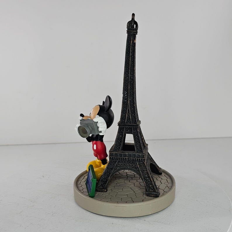 K&ouml;nnte beinhalten: Eine Figur von Mickey Mouse mit einer Kamera, neben einer schwarzen Nachbildung des Eiffelturms. Der Sockel ist hellgrau mit einem Kopfsteinpflastermuster. Mickey Mouse tr&auml;gt rote Shorts, gelbe Schuhe und ein wei&szlig;es Hemd. Der Turm ist etwa 30 cm hoch.