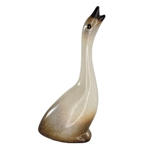 Puede incluir: Una figura de cerámica de un ganso con el cuello largo y esbelto extendido hacia arriba y el pico abierto. El cuerpo presenta un acabado liso y brillante con un degradado de color, pasando de un crema claro en la parte superior a un marrón oscuro.