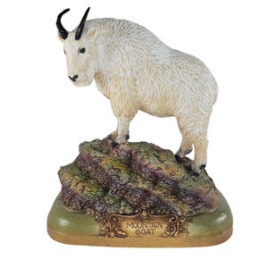 Può includere: Statuetta dettagliata di un camoscio in piedi su una base rocciosa. La capra ha un mantello bianco strutturato, corna nere e un viso dettagliato. La base è multicolore con toni verdi, marroni e dorati e presenta la scritta "MOUNTAIN GOAT".