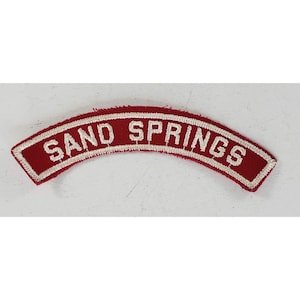 May include: A red, arched patch with white embroidered text reading "SAND SPRINGS". The patch has a white border and a textured appearance. The text is in a sans-serif font. This patch is likely a collectible or souvenir.