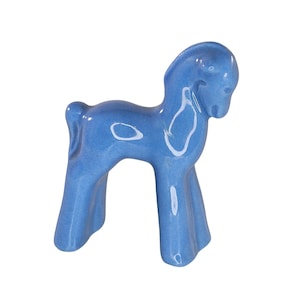 Puede incluir: Pequeña figura de caballo de cerámica azul claro. El caballo tiene un diseño estilizado con un cuerpo curvo y patas acampanadas. La figura es brillante y parece ser un objeto decorativo, posiblemente para una estantería o un escritorio.