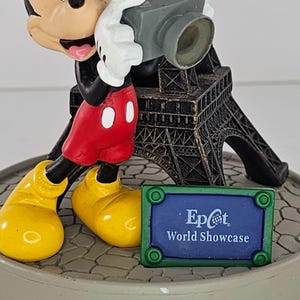 K&ouml;nnte beinhalten: Eine Mickey Mouse-Figur, die eine Kamera h&auml;lt, mit einem Miniatur-Eiffelturm im Hintergrund. Auf dem Sockel steht "Epcot World Showcase". Mickey tr&auml;gt rote Shorts, gelbe Schuhe und wei&szlig;e Handschuhe.
