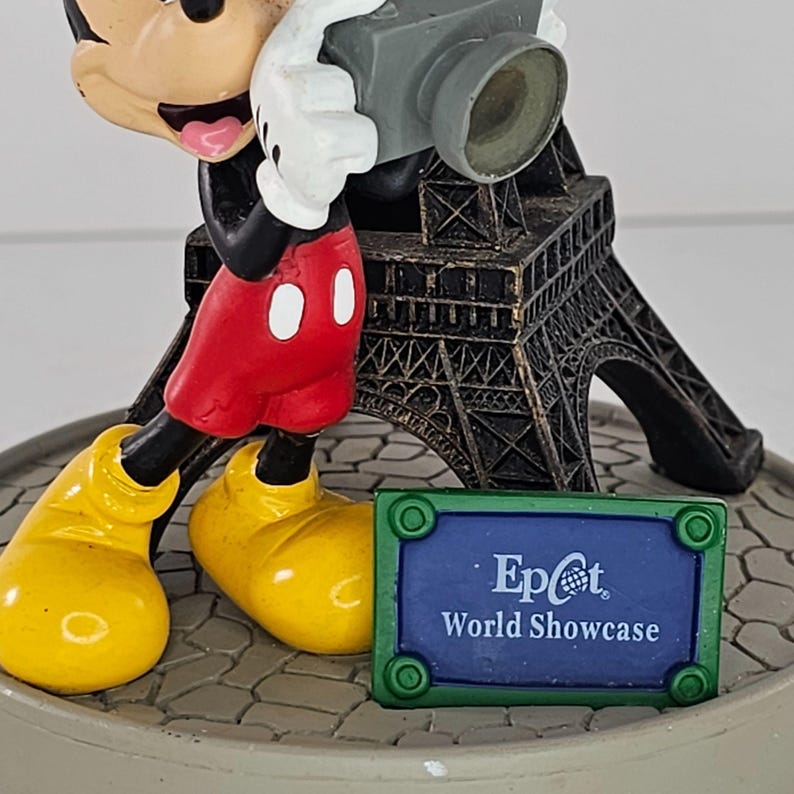 K&ouml;nnte beinhalten: Eine Mickey Mouse-Figur, die eine Kamera h&auml;lt, mit einem Miniatur-Eiffelturm im Hintergrund. Auf dem Sockel steht "Epcot World Showcase". Mickey tr&auml;gt rote Shorts, gelbe Schuhe und wei&szlig;e Handschuhe.