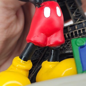 K&ouml;nnte beinhalten: Eine Figur mit Mickey Mouse's roten Shorts mit wei&szlig;en Kn&ouml;pfen, schwarzen Beinen und gelben Schuhen. Die Figur steht vor einer Miniatur des Eiffelturms und einem gr&uuml;nen und blauen Element.