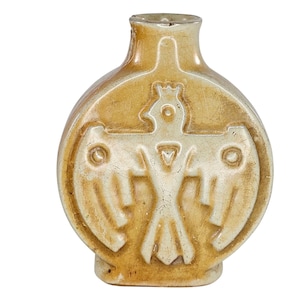 May include: A small, antique-style ceramic bottle in tan and off-white. The bottle features a raised relief design of a stylized bird with outstretched wings. It has a rounded body and a narrow neck. The bottle is approximately 10 cm tall.