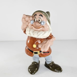 Könnte beinhalten: Eine Keramikfigur von Doc aus Schneewittchen und den sieben Zwergen. Doc trägt einen braunen Mantel, einen grünen Hut und eine runde Brille. Die Figur hat einen weißen Bart und hält die Hand an den Kopf. Ein Aufkleber auf der Figur lautet "DOC AMERICAN ARTISTS LOS ANGELES."