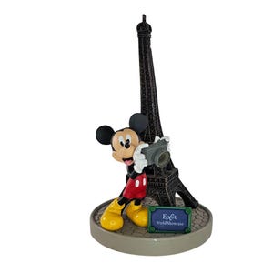 K&ouml;nnte beinhalten: Eine Mickey Mouse-Figur, die eine Kamera vor einer schwarzen Nachbildung des Eiffelturms h&auml;lt. Mickey tr&auml;gt rote Shorts, gelbe Schuhe und wei&szlig;e Handschuhe. Der Sockel tr&auml;gt ein Schild mit der Aufschrift "Epcot World Showcase".