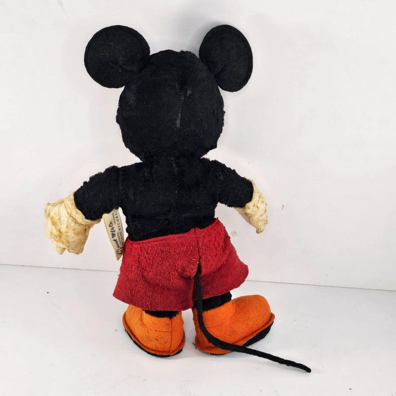 May include: A vintage plush Mickey Mouse toy with black fur, red shorts, and orange shoes. The toy has a tag that reads "Disney Mickey Mouse".