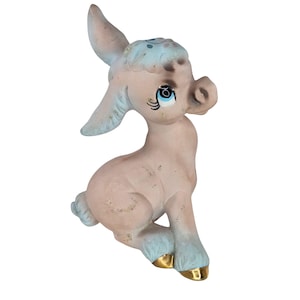 Puede incluir: Figura de burro de cerámica vintage en posición sentada. El burro tiene un cuerpo rosa claro con detalles azules en las orejas, la cabeza y las pezuñas. Las pezuñas están adornadas con dorado. El burro tiene grandes ojos azules y una expresión dulce.