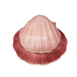 May include: A ceramic lidded dish shaped like a seashell with a glossy finish. The top lid is a pale, dusty pink with vertical ridges, while the base plate is a darker, muted rose colour with a scalloped edge and matching vertical ribbing.