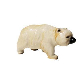 May include: A ceramic polar bear figurine with a glossy, off-white crackle glaze finish. The bear is depicted in a walking stance, featuring a smooth, rounded body, small ears, and a distinct, shiny black nose that contrasts with the light body.