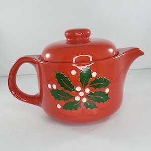 May include: Red ceramic teapot with a green holly leaf and white berry design. The teapot has a lid and a handle.