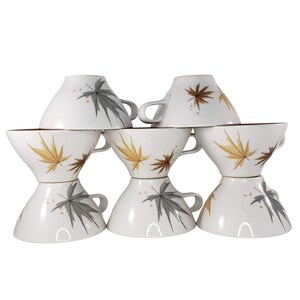 May include: A set of six white ceramic cups arranged in three stacks of two. Each cup features a pattern of scattered maple leaves in shades of brown, gold, and grey. The cups have a wide, flared rim and a small handle on the side.