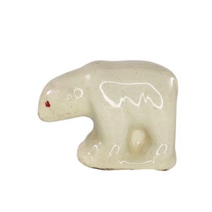 May include: A small, glossy, off-white ceramic figurine of a polar bear in a walking pose. The bear has a smooth, reflective surface with subtle speckling and a single, small, distinct red dot painted on the side of its snout near the nose.
