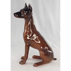 May include: A brown ceramic Doberman Pinscher figurine in a seated position. The dog has a glossy finish with a dark brown head and ears. The figurine is detailed with white accents and sits on a white surface.