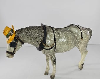 May include: A dapple gray horse figurine wearing a black leather harness with gold-toned chain accents and a small yellow straw hat with a green band perched on its head. The horse is depicted in a standing position with a downward-facing head.