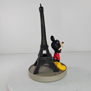 K&ouml;nnte beinhalten: Dekorative Figur mit einem Miniatur-Eiffelturm und einer Mickey Mouse-Figur. Der Eiffelturm ist dunkelbraun, und Mickey Mouse tr&auml;gt rote Shorts und gelbe Schuhe. Der Sockel ist hellgrau.