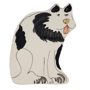 May include: A ceramic dog figurine in a sitting position. The dog is primarily white with black markings on its ears, back, and a spot on its side. The dog's face features a brown nose, a red tongue, and a whimsical expression.