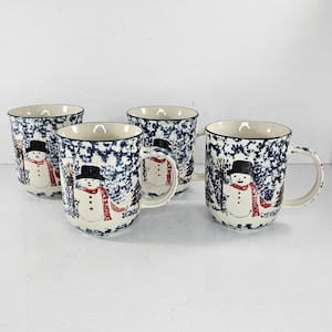 Op de afbeelding: Vier keramische mokken met een winterthema. Elke mok heeft een sneeuwpopontwerp met een zwarte hoge hoed, rode sjaal en een bezem. De mokken hebben een gespikkeld blauw-wit patroon en een donkerblauwe rand.