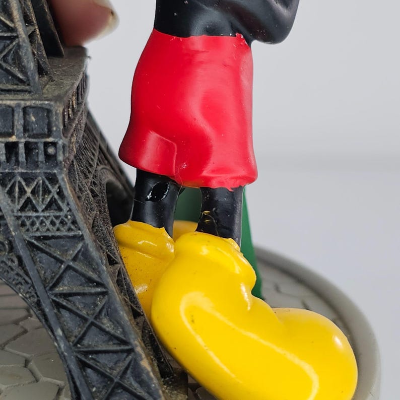 K&ouml;nnte beinhalten: Eine Figur einer Zeichentrickfigur mit rotem Rock, schwarzen Beinen und gelben Schuhen, die neben einem Miniatur-Eiffelturm steht. Die Figur steht auf einem kreisf&ouml;rmigen Sockel mit grauem und wei&szlig;em Design. Der Eiffelturm ist grau.
