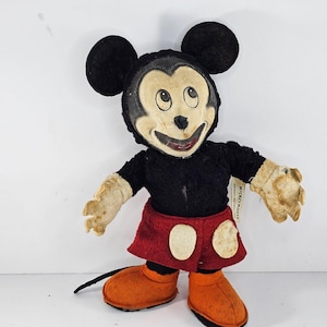 May include: Vintage Mickey Mouse plush toy with a black and red outfit. The toy has a worn and faded appearance, with a tag that reads "Mickey Mouse".