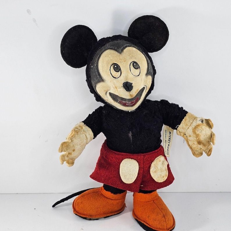 May include: Vintage Mickey Mouse plush toy with a black and red outfit. The toy has a worn and faded appearance, with a tag that reads "Mickey Mouse".