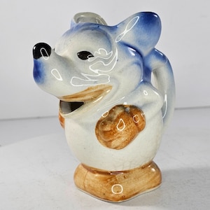 Puede incluir: Una jarra de cerámica con forma de ratón de dibujos animados con detalles pintados en azul y blanco. El ratón sostiene un objeto marrón en su mano. La jarra tiene una base marrón.
