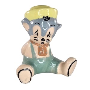 May include: A vintage ceramic figurine of a seated cartoon cat wearing a yellow hat and blue overalls. The cat has a light blue face, pink ears and arms, and black accents. The figurine is a collectible item.