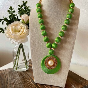 May include: A green silk thread necklace with gold beads and a large green circular pendant with a pearl in the center. Matching green silk thread earrings with gold beads are also included.