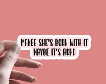 May include: White sticker with black text that reads "Maybe she's born with it maybe it's ADHD". The sticker is being held by a hand against a pink background.