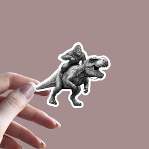 May include: A black and white sticker of a gorilla riding a Tyrannosaurus Rex. The sticker is on a light brown background.