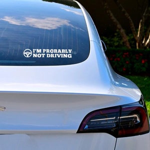 May include: A white Tesla Model 3 with a rear window sticker that reads "I'm probably not driving".
