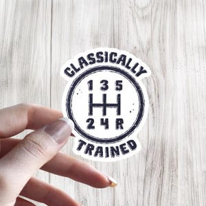May include: A white sticker with a black gear shift design and the words "Classically Trained". The gear shift pattern shows 1, 3, 5, 2, 4, and R. The sticker is held by a hand against a light wood background.