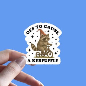 May include: A white sticker with a raccoon riding a bicycle, wearing a party hat, and the text "Off to cause a kerfuffle." The sticker has a blue background and the shop's logo in the bottom right corner.